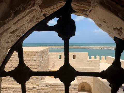 Sea view framed by a stone window of an old fortress looking toward turquoise waters and breakwater.