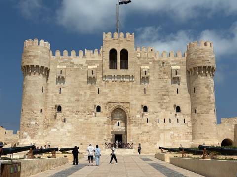 Front façade of Qaitbay Citadel with cannons and tourists under a bright blue sky.