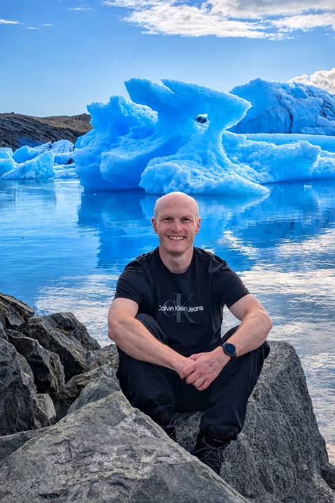 Smiling traveller seated on rocks beside vivid blue glacial lagoon dotted with floating icebergs.