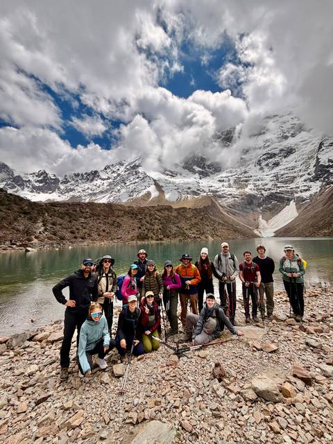 Hiking group posing beside a turquoise alpine lake backed by snowy Andean peaks and dramatic clouds.