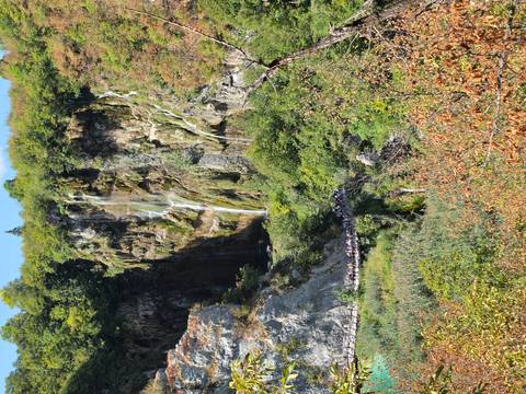 Tall waterfall drops down a rocky cliff above a wooden footbridge crowded with visitors.