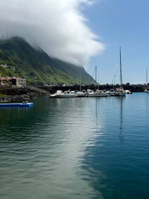 Quiet marina with sailboats moored below lush green cliffs partially wrapped in cloud.