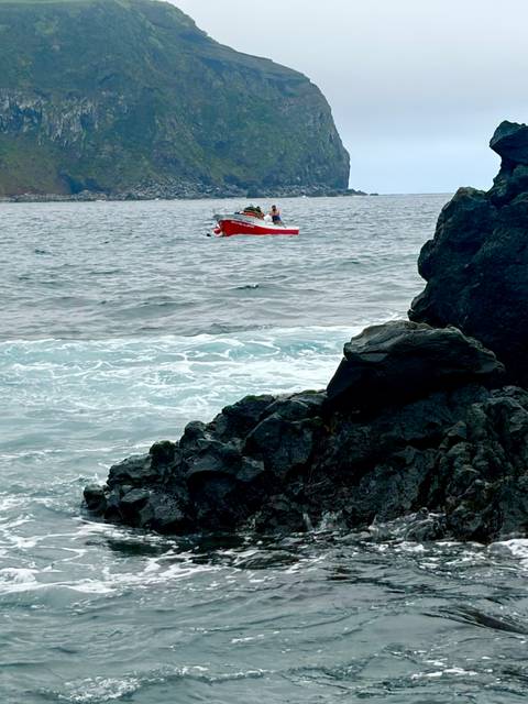Small red fishing boat on choppy sea near dark volcanic rocks.