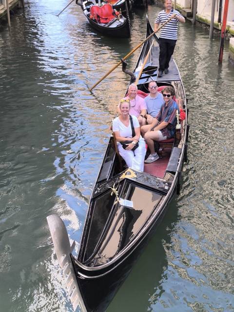 Four travellers sit in a classic black gondola gliding through a narrow Venetian canal with rippling water all around.
