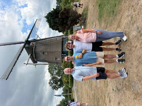 A family stands in front of a traditional thatched windmill on a grassy Dutch landscape under a partly cloudy sky.