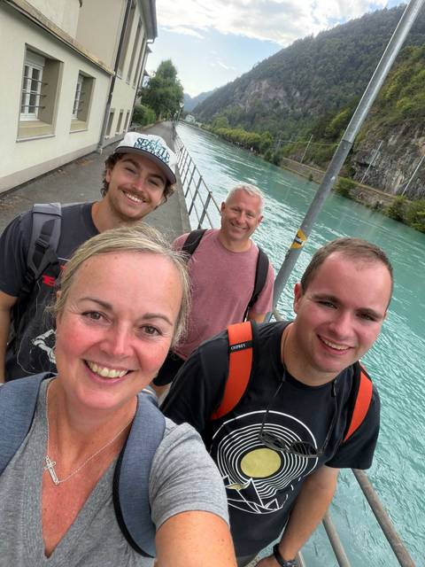 A close-up selfie of four smiling travellers walking beside a bright turquoise river with rocky banks.