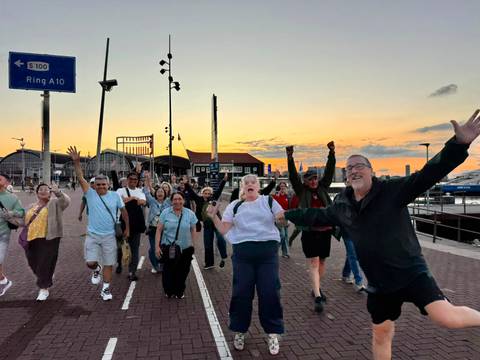 Energetic group of travelers celebrates while walking along a harbor promenade at sunset.
