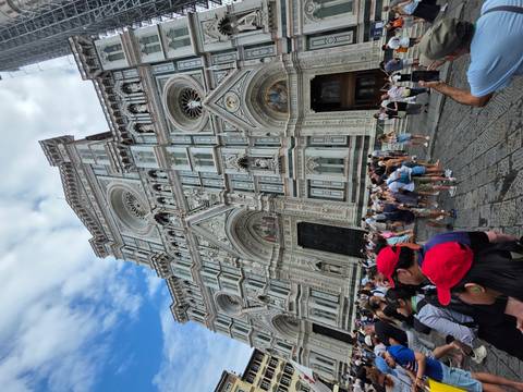 Tourists milling before the multicolored marble facade of Florence’s Duomo under a partly cloudy sky.