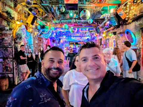 Two men taking a selfie inside a vibrant, neon-lit bar filled with people