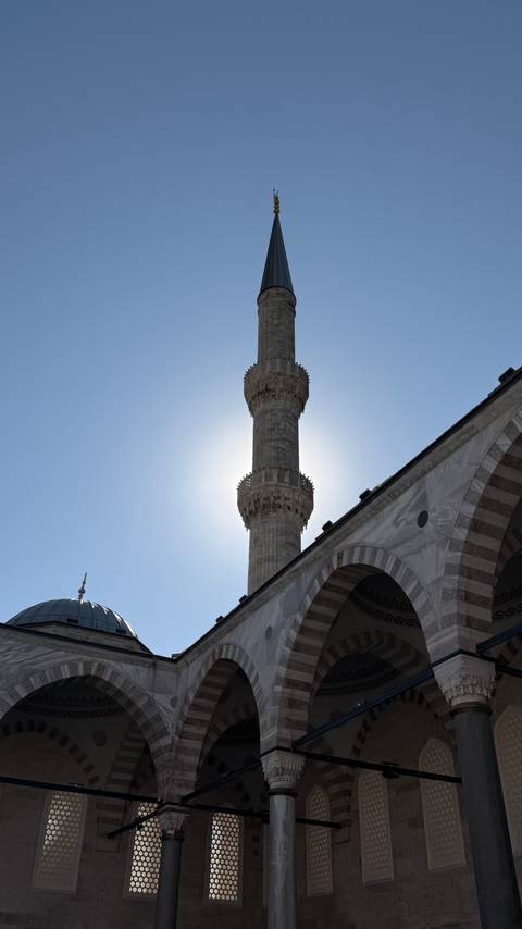 Tall stone minaret of a Turkish mosque silhouetted against the sun with clear blue sky.