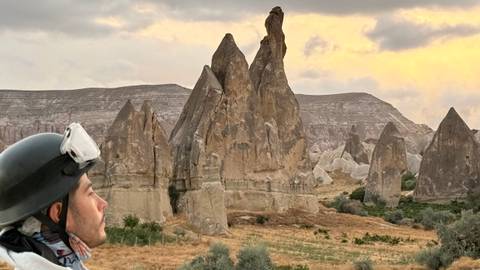 A traveler wearing a helmet gazes at dramatic fairy-chimney rock formations under a glowing evening sky in Cappadocia.