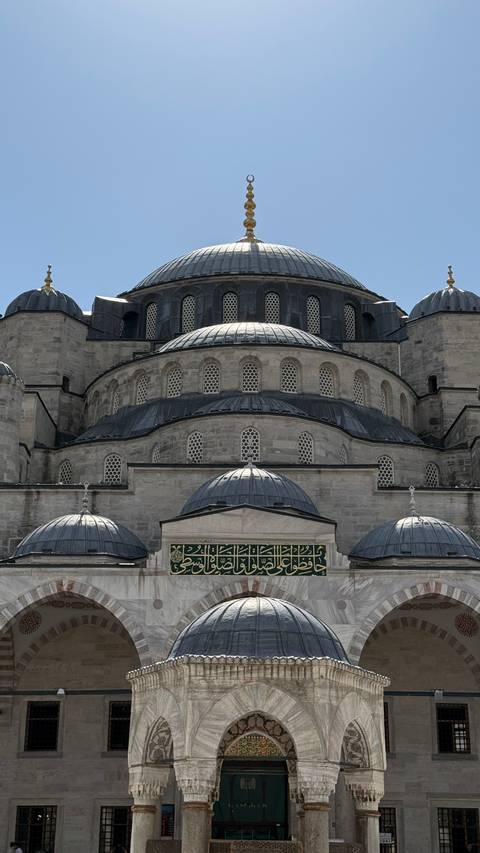 Close view of grey stone domes and stacked arches of a grand Ottoman mosque against a clear sky.