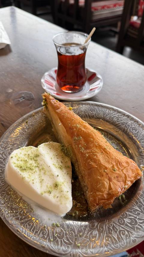 Silver plate with a slice of pistachio-topped baklava and vanilla ice cream beside a tea glass.