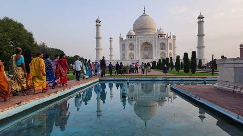 Visitors admire the Taj Mahal reflected perfectly in a blue pool during the warm glow of sunset.