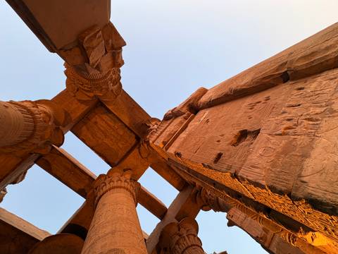Upward view of sandstone columns and carved beams glowing in warm evening light against a pale sky.