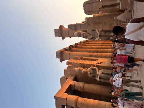 Crowds explore an ancient temple courtyard lined with giant seated pharaoh statues at dusk.