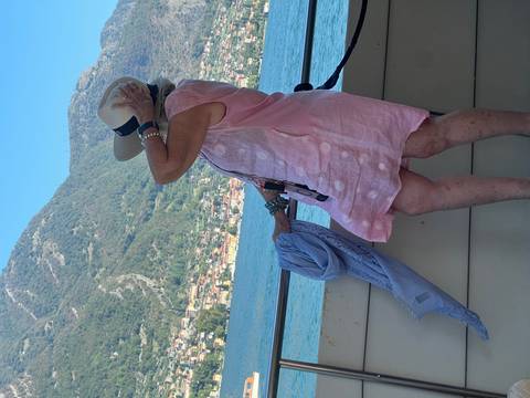Older woman in pink dress holding her hat on a lakeside boat with mountains behind
