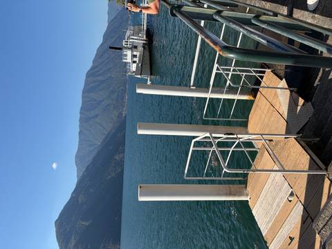 Pier with white mooring posts extending into a deep blue alpine lake with ferry boat and mountains