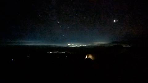 Low-light night scene of a distant town’s lights and starry sky viewed from a dark mountainside camp.