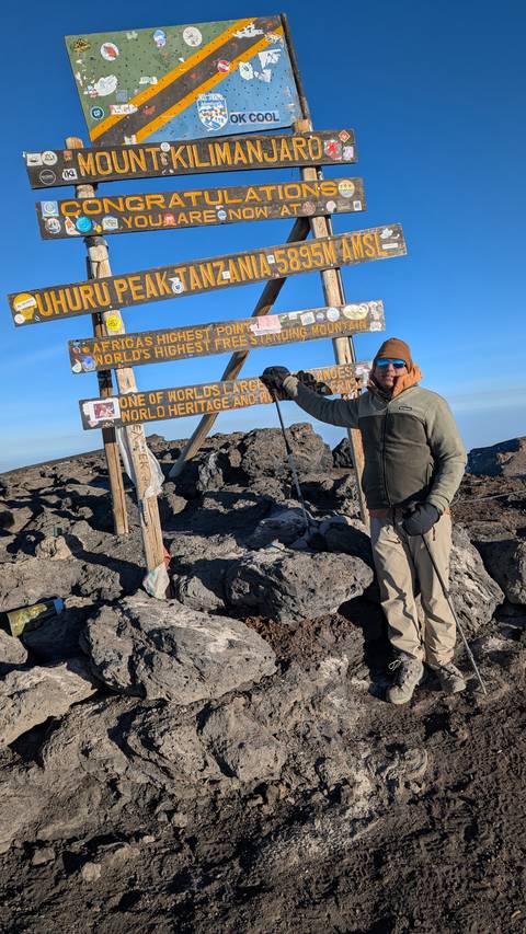 Climber in winter gear posing beside the Uhuru Peak sign atop Mount Kilimanjaro.