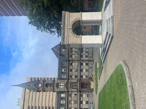 Historic sandstone civic building with clock tower beside a stone arch monument in a city square.