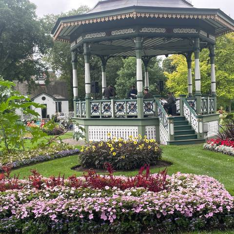 Musicians perform in a Victorian bandstand surrounded by colorful flowerbeds in a public garden.
