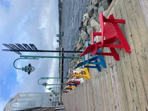 Colorful Adirondack chairs line a wooden boardwalk overlooking a harbor under moody skies.