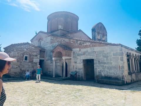 Tourists explore an old stone church with Byzantine architecture under a bright blue sky.