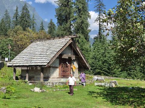 Small stone cottage surrounded by lush conifer forest with travellers nearby