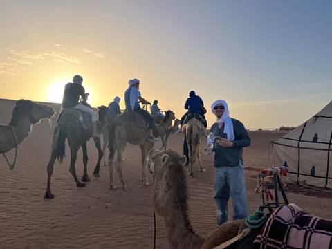 Travellers ride camels across desert dunes with the sun setting behind them near a Berber tent.