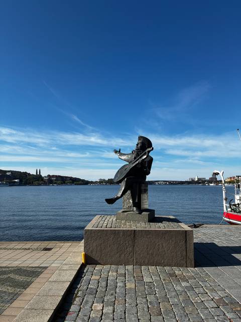 Waterfront bronze statue of a musician gestures toward Stockholm’s broad harbor under blue skies.