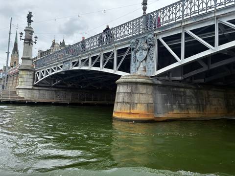 Ornate steel bridge spans green canal waters with decorative crest and city buildings above.