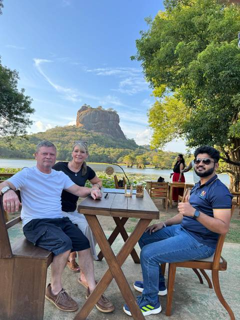 Travellers relax at an outdoor café beside a lake with the iconic Sigiriya Rock rising in the background.