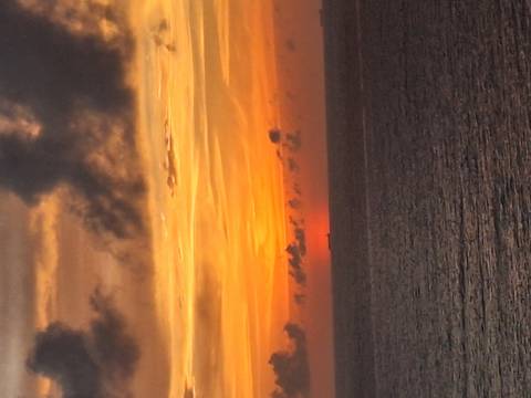Fiery orange and gold sunset over a calm ocean with a distant ship on the horizon and dramatic clouds overhead.
