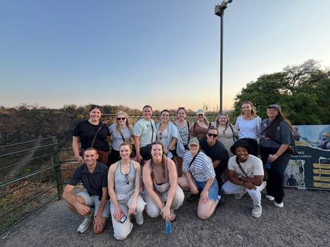 A smiling tour group poses at a scenic viewpoint with a deep gorge and bridge in the background.