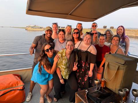 Tour group on a river boat safari poses for a photo with elephants grazing on the riverbank behind.