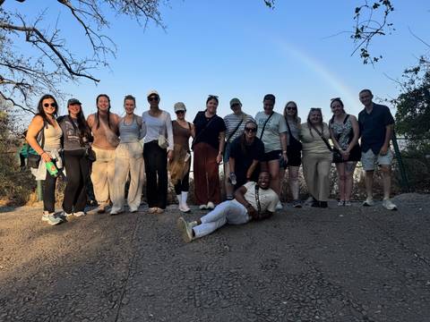 Large hiking group stands on a dirt road with a faint rainbow arching across the clear sky.