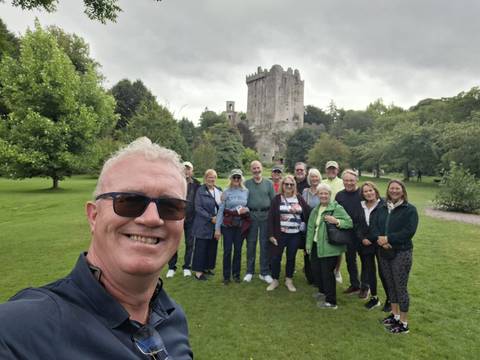 Tour group selfie on green lawn in front of stone Blarney Castle on cloudy day.