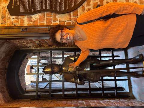 Visitor smiles beside a suit of armor inside a brick-walled medieval hall.