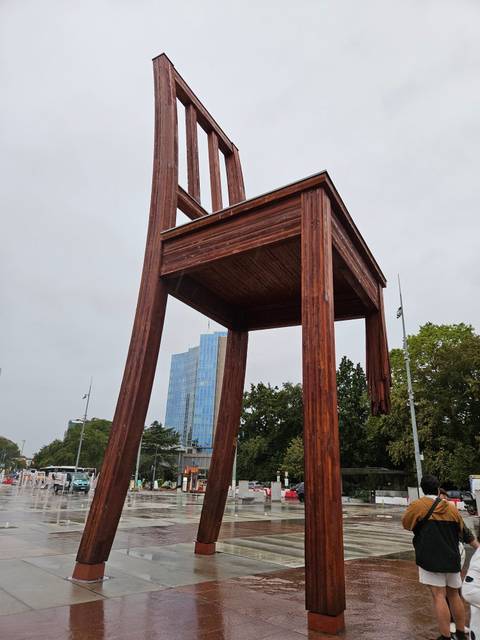 Giant wooden 'Broken Chair' sculpture stands tall near modern glass UN building against a grey sky