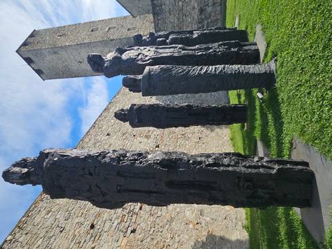 Row of tall, textured black sculptures stand beside an ancient stone wall and tower