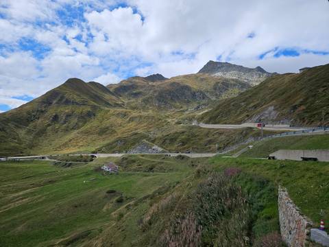 Mountain road winds across alpine pastures beneath rugged green and brown peaks under patched clouds