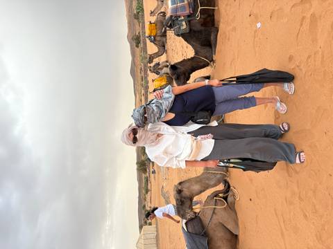 Two women wearing desert scarves pose beside seated camels on orange sand with low hills and cloudy skies in the background.