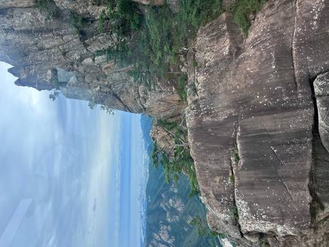 Towering jagged rock face rises above forested mountains with a distant coastal city visible under a cloudy sky.