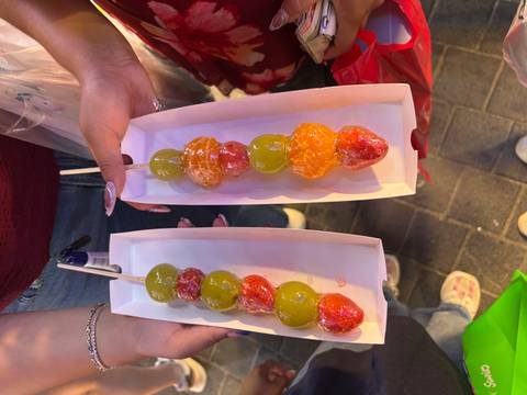 Two trays of colorful candied fruit skewers held up at a bustling night market.