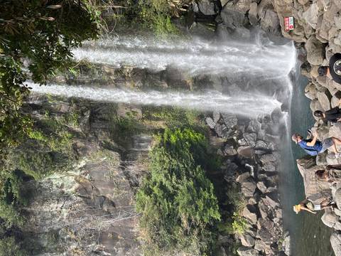 Visitors photograph a tall waterfall plunging into a rocky pool amid lush greenery.