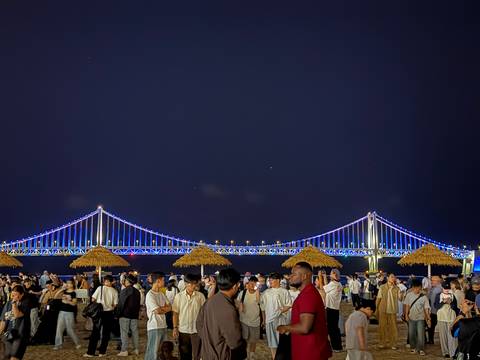 Nighttime beach crowd under straw parasols watching the brightly lit Gwangan Bridge span the dark sky.