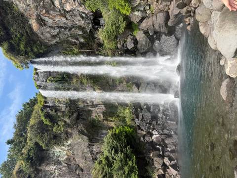 Twin waterfalls cascade down a moss-covered cliff into an emerald pool on a sunny day.