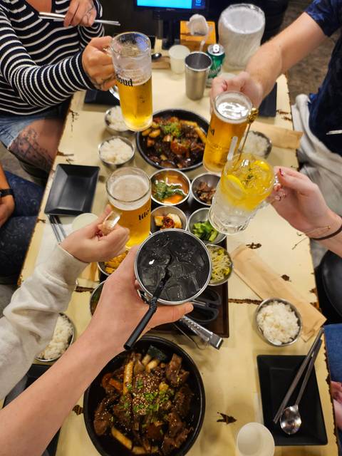 Overhead view of diners clinking varied drinks above a Korean feast of shared side dishes.