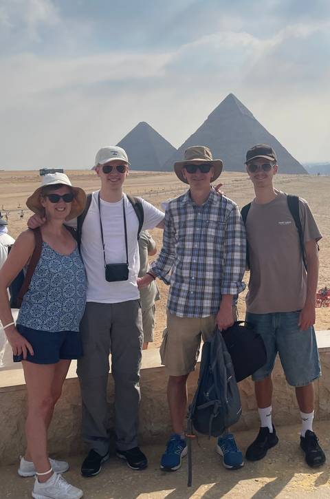 Family of four posing with desert landscape and one of the Giza pyramids faintly visible in the background on a sunny day.